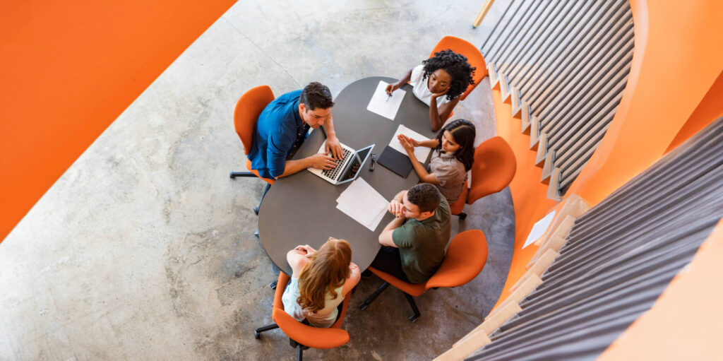 Five people sit around an oval table with a laptop, papers, and tablet, viewed from above. The chairs are orange, and the room has curved, ribbed walls, creating a modern workspace ambience.