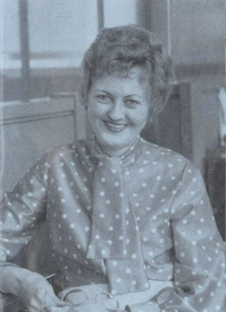 Black and white photo of a woman smiling, wearing a polka dot blouse with a tie-neck. She is holding a pair of glasses in one hand. The background features office furniture, including a desk and shelves.