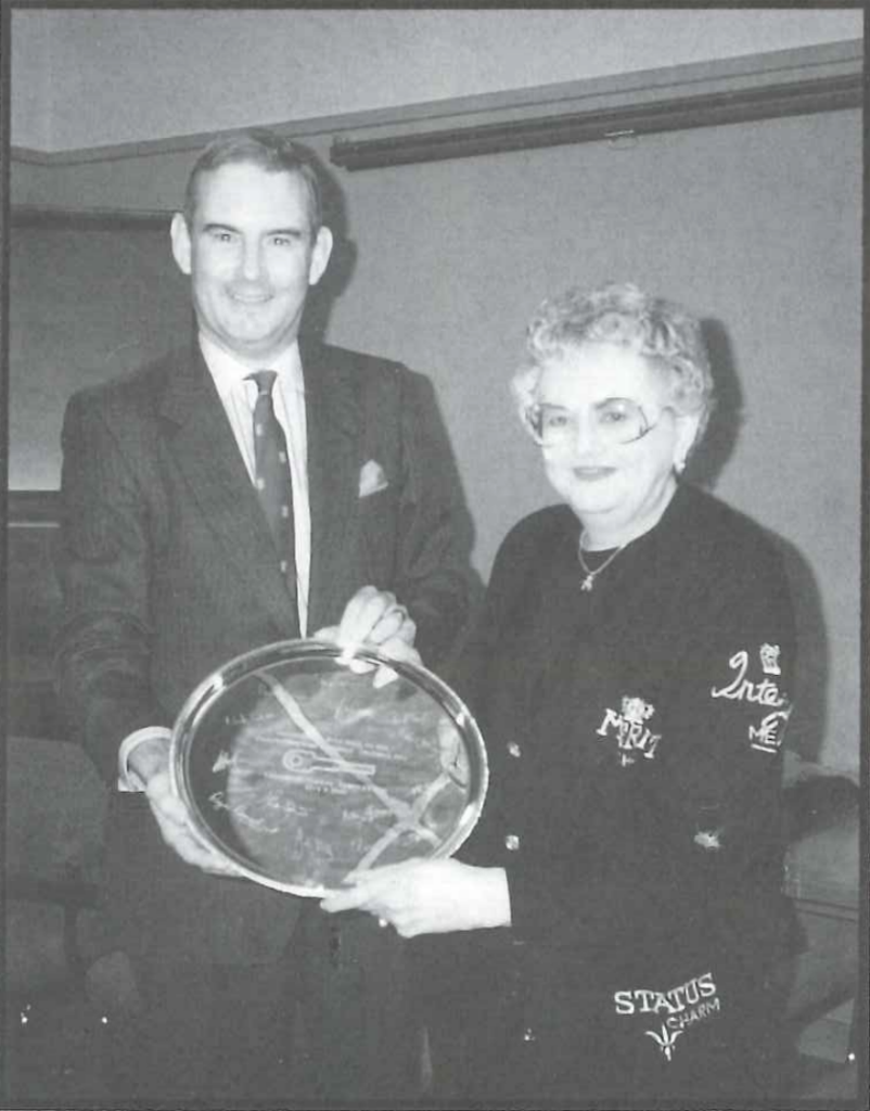 A man and a woman standing together, both smiling. They are holding a large circular award or platter with text and signatures. The man is wearing a suit, and the woman is in a decorated sweater. They appear to be at an indoor event or ceremony.