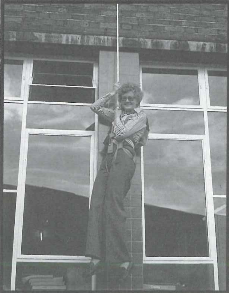 A person dressed in 1970s-style clothing hangs from a rope in front of a large window on a brick building, appearing to playfully dangle or pretend to climb.