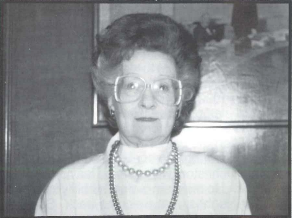 A grayscale photo of an older person with short, voluminous hair and large glasses. They are wearing a light-colored top and a long, beaded necklace, standing in front of a framed picture.