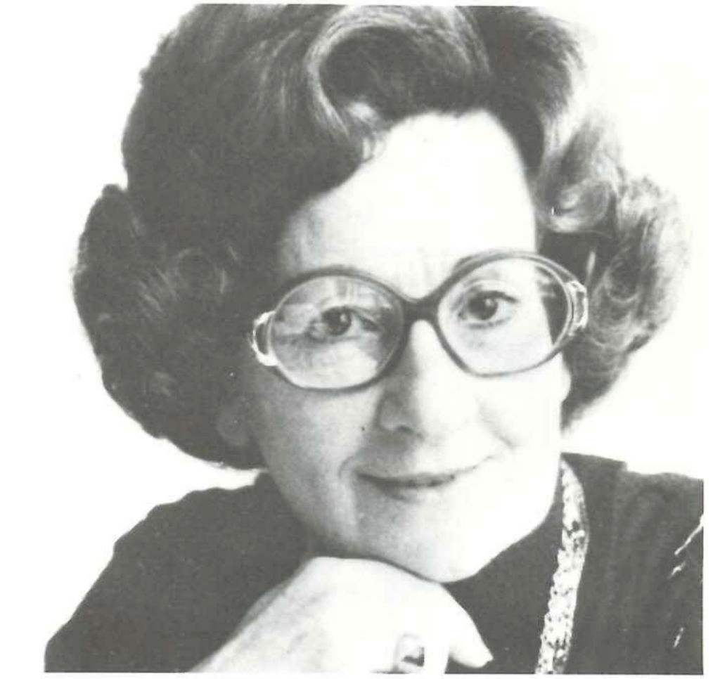 A woman with curly hair and large round glasses is seen in a black-and-white portrait. She rests her chin on her hand and wears a dark top with a chain necklace.