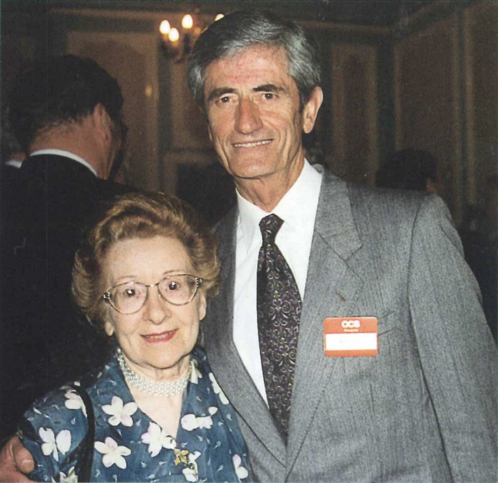 An elderly woman in a floral dress and glasses smiles while standing next to an older man in a gray suit and tie. They are indoors at a formal event. The man has a red name tag on his suit.