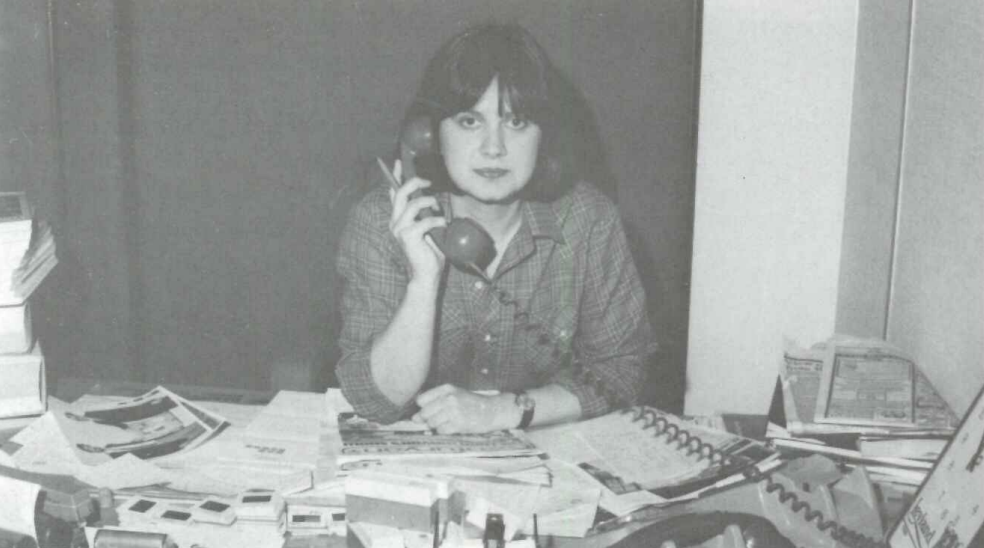 A person with shoulder-length hair is sitting at a cluttered desk, holding a phone up to their ear. The desk is filled with papers, books, and office supplies. The image is in black and white.