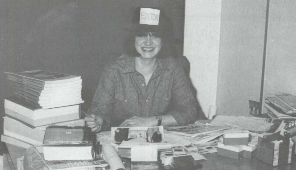 A person wearing a paper hat labeled Editor sits at a cluttered desk filled with books, papers, and office supplies. They are smiling and holding a pencil. The setting appears to be an office. The image is in black and white.