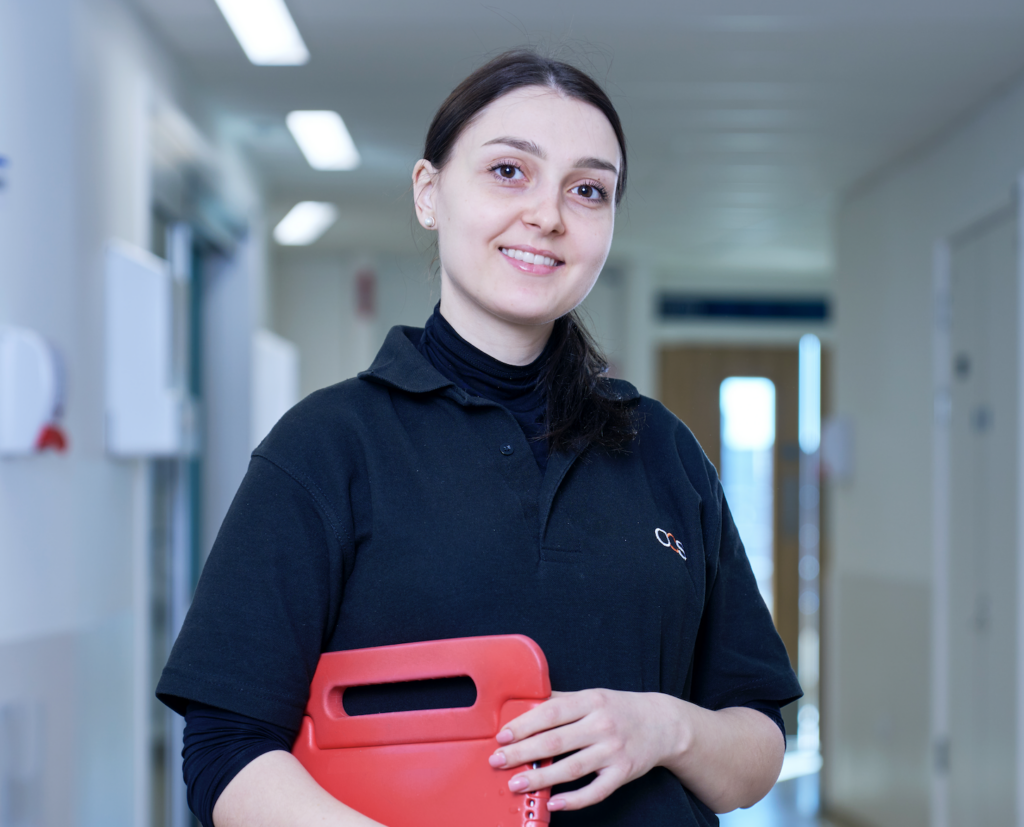 A woman wearing a black polo shirt stands in a hallway, smiling and holding a red plastic folder. The background shows a well-lit corridor with white walls and doors.