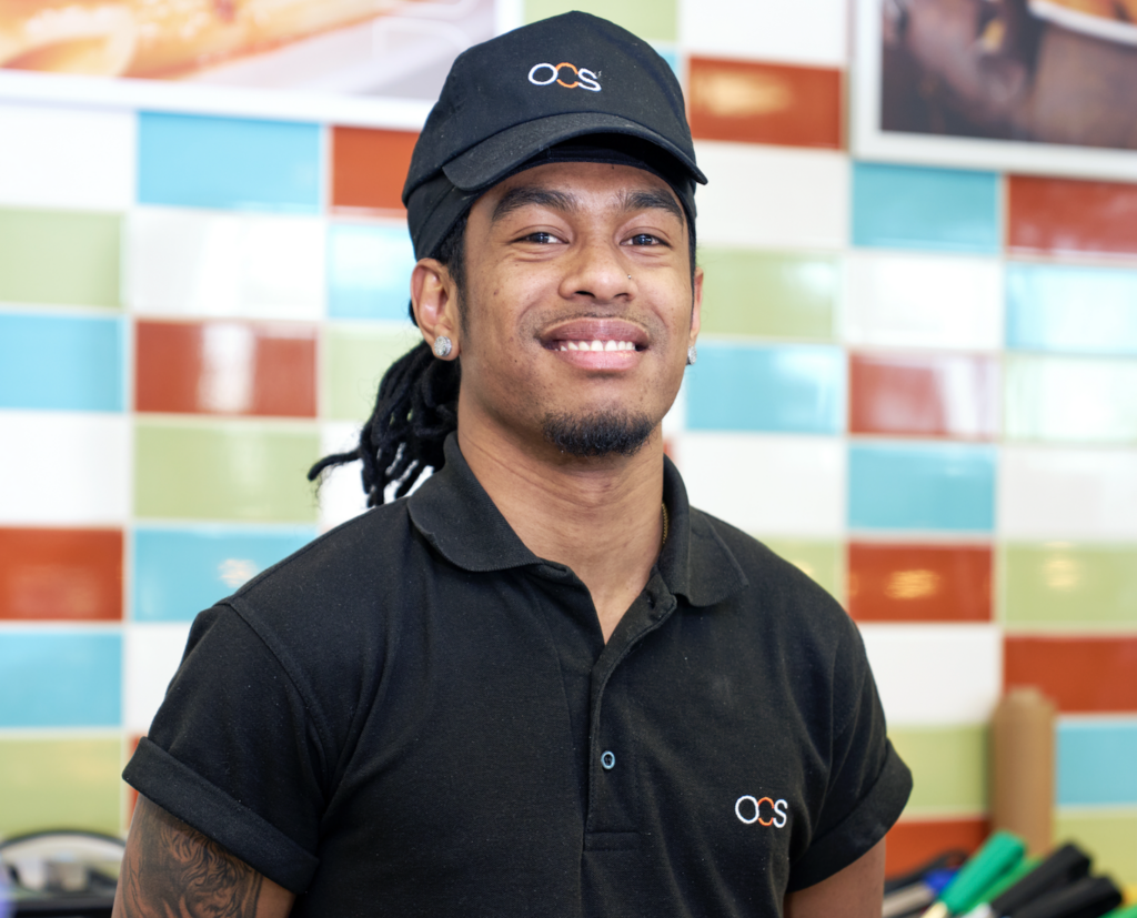 A smiling person wearing a black OCS uniform and cap stands in front of a colorful tiled wall in what appears to be a restaurant or café.