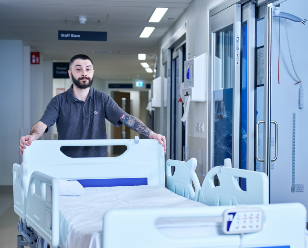 A man with a beard and tattoos on his arm pushes an empty hospital bed down a brightly lit hospital corridor near a sign that reads Staff Base.