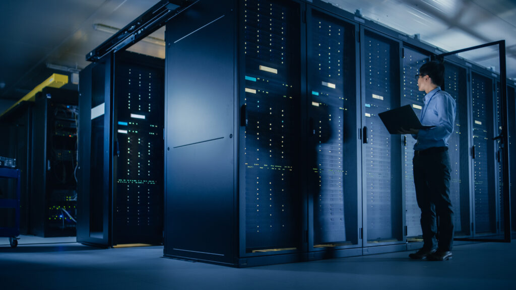 A person holding a laptop stands in a dark, modern server room, examining tall racks filled with servers and blinking indicator lights.