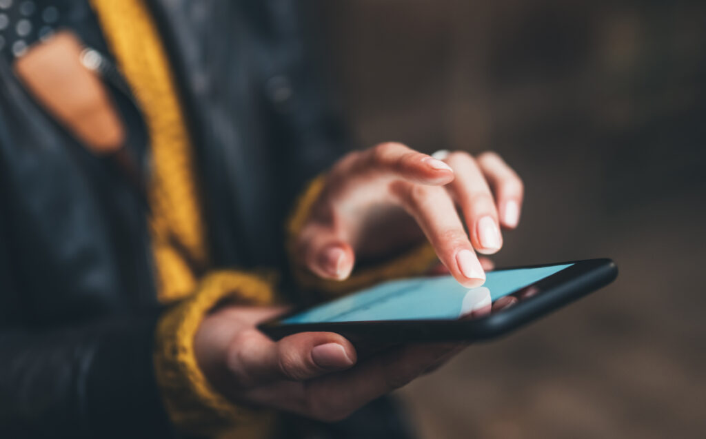 A person wearing a black jacket and yellow sweater uses their index finger to navigate on a smartphone, focusing on their hands and the device.