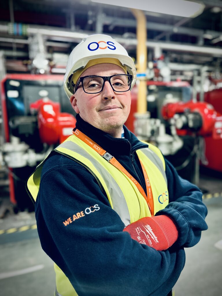 A man wearing glasses, a white hard hat, a high-visibility vest, and gloves stands with arms crossed in an industrial setting. He has an OCS logo on his helmet, vest, and lanyard. Red machinery is visible in the background.