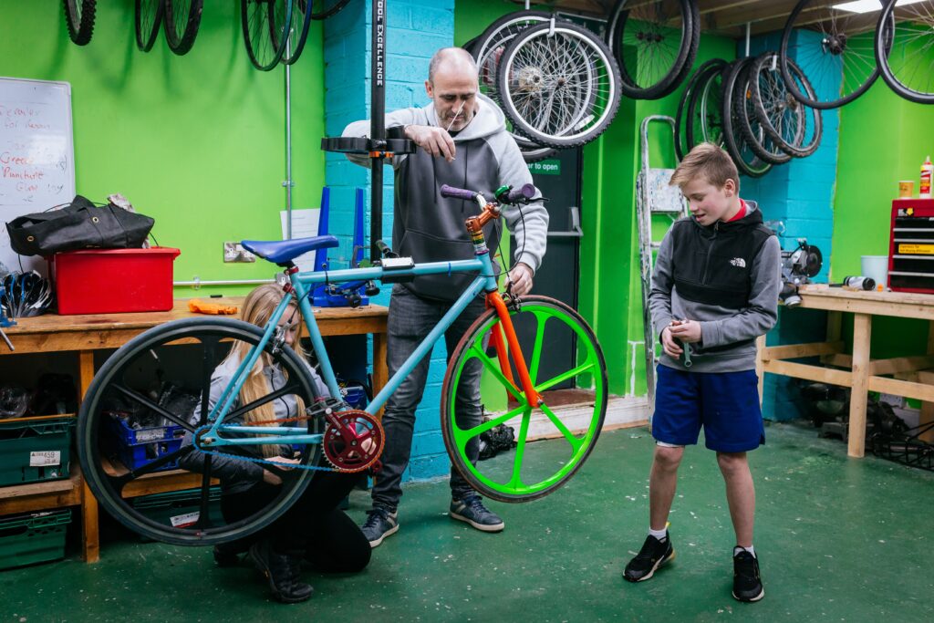 Two people work on a colorful bicycle in a workshop, while a boy stands nearby watching. The workshop has green walls, tools, and bicycle wheels hanging on the wall.
