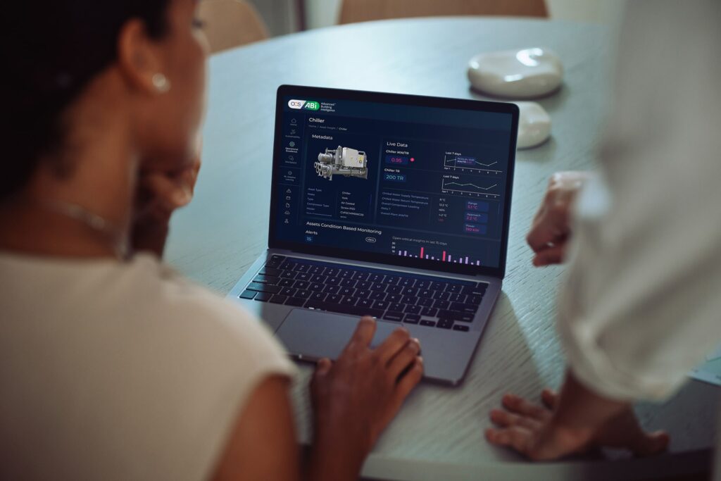 Two people look at a laptop displaying a dashboard with equipment data, graphs, and charts. The laptop is on a round table and their faces are not visible.