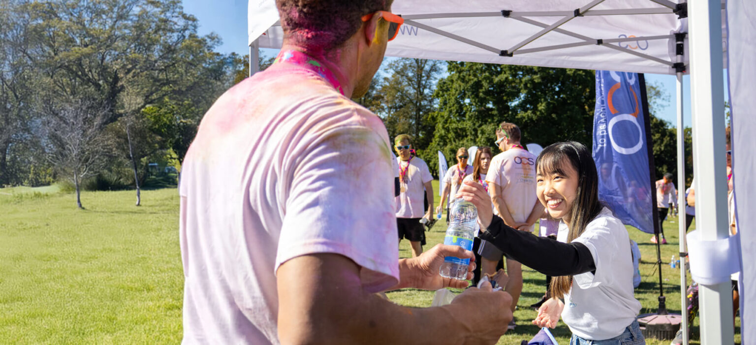 A smiling woman hands a water bottle to a man covered in colored powder under a tent at an outdoor event, with other participants and trees visible in the background.