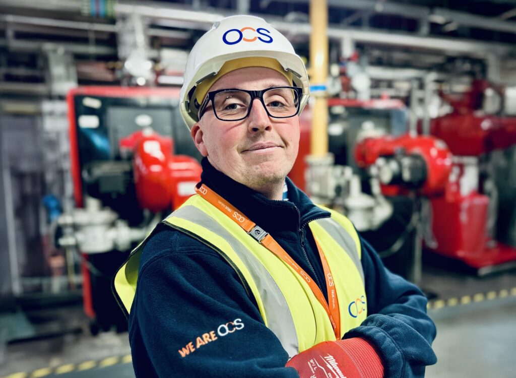 A person wearing safety glasses, a white hard hat, and a high-visibility vest stands with arms crossed in an industrial setting. They have an OCS logo on their hat, vest, and lanyard. Machinery is visible in the background.