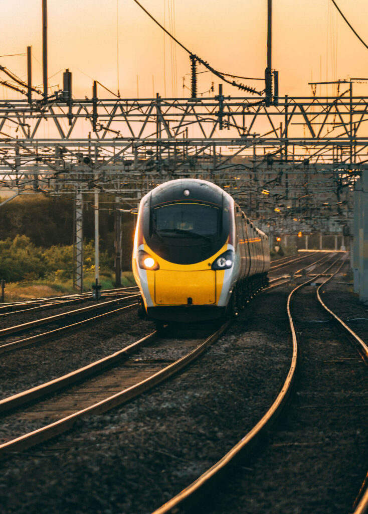 A modern yellow and black train travels on curved tracks under a network of overhead wires at sunset, with greenery visible to the left and a warm orange sky in the background.