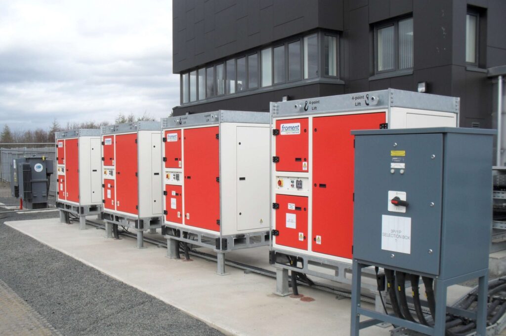 A row of industrial electrical control panels with red and white doors stands outside a modern building, connected by metal conduits and cables, on a concrete platform.