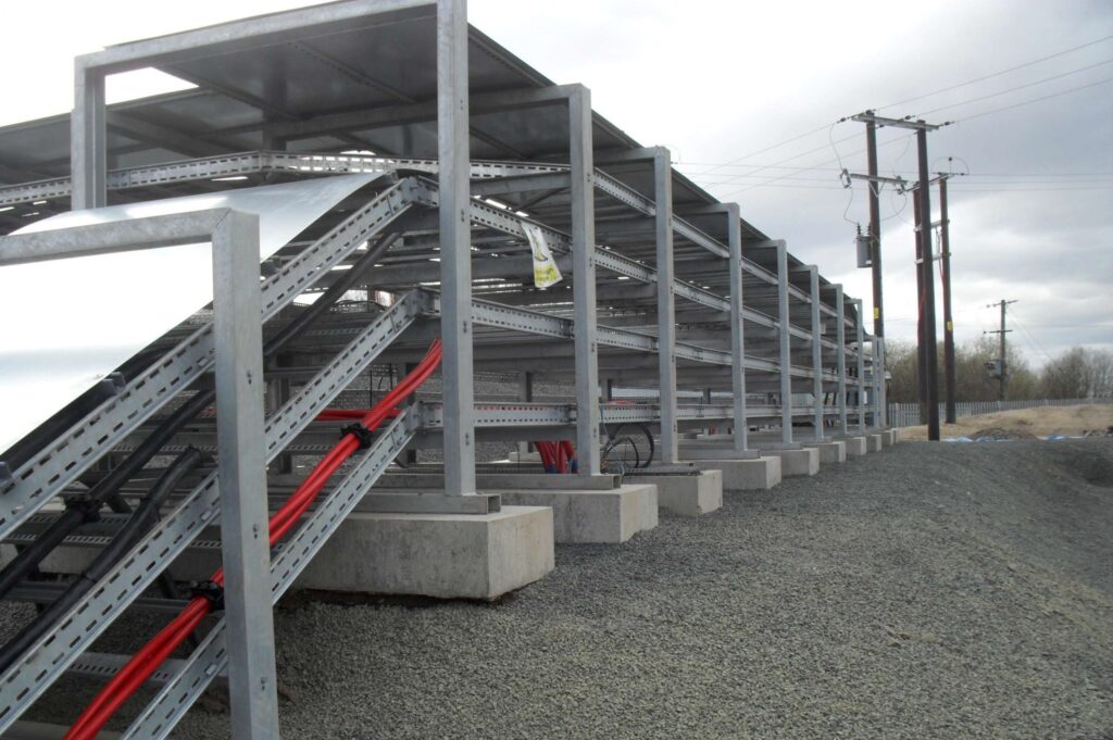 Large metal cable trays supported by concrete blocks run along a gravel path outdoors, with red and black cables inside. Overhead, there is a metal roof structure, and power poles are visible in the background under a cloudy sky.
