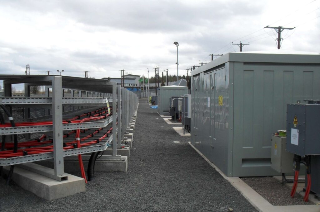 Outdoor energy facility with electrical equipment, large cabinets, and exposed cables on racks, set on gravel grounds with utility poles and fencing in the background under a cloudy sky.