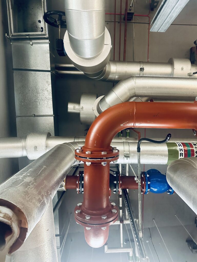 Industrial pipes and ducts on a ceiling, featuring a prominent red pipe with bolts, surrounded by silver ventilation ducts and other machinery components. The perspective is looking upwards.