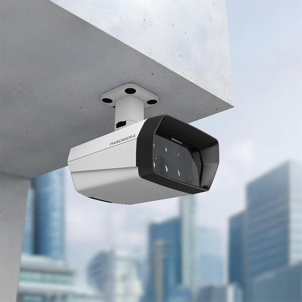 A modern security camera is mounted on a concrete ceiling outdoors, with a blurred cityscape of tall buildings in the background.