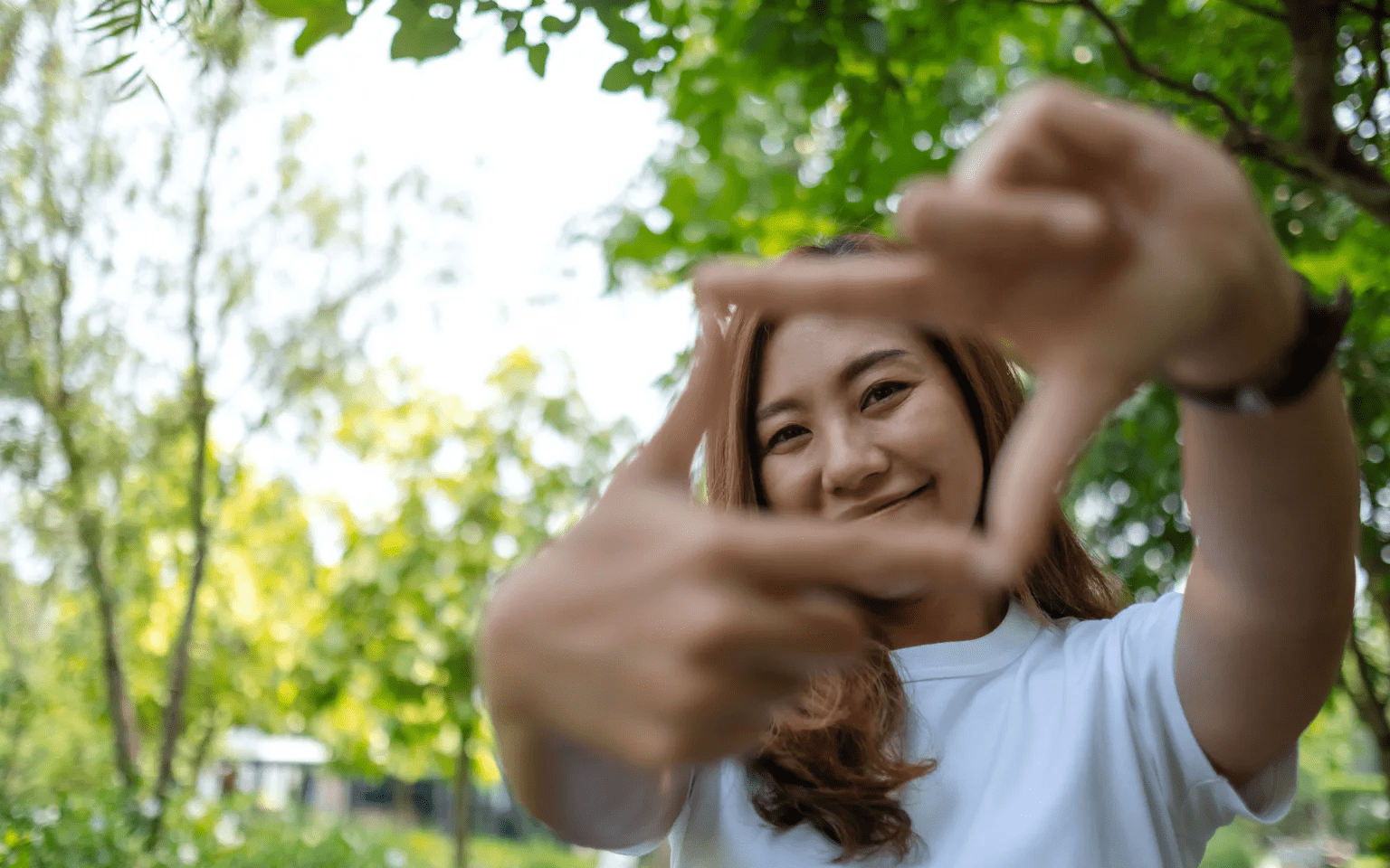 A woman in a white shirt smiles outdoors, holding her hands up to frame her face with her fingers, with green trees and sunlight in the background.