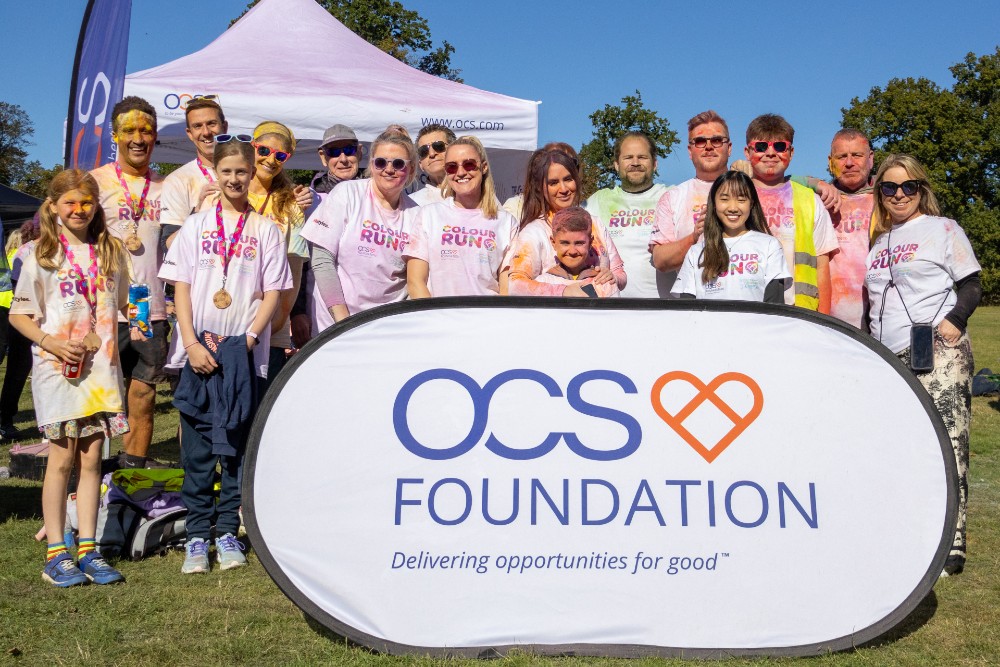 A group of people wearing white OCS Colour Run shirts, some with colored powder on their clothes and faces, stand smiling behind an OCS Foundation banner at an outdoor event.