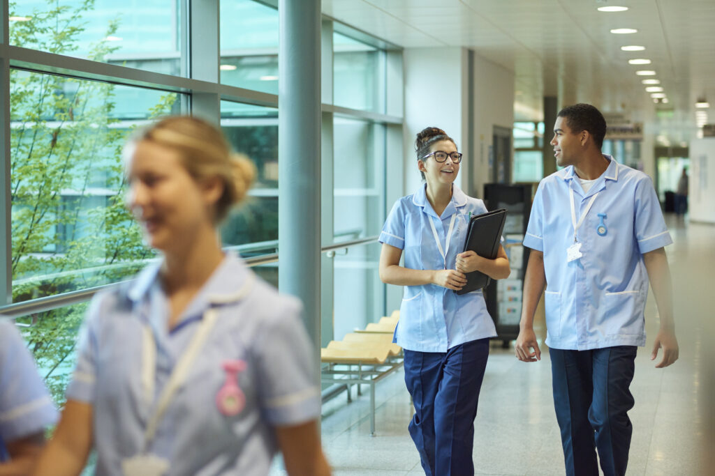 A group of healthcare professionals in light blue uniforms walk and talk together in a bright hospital corridor with large windows.