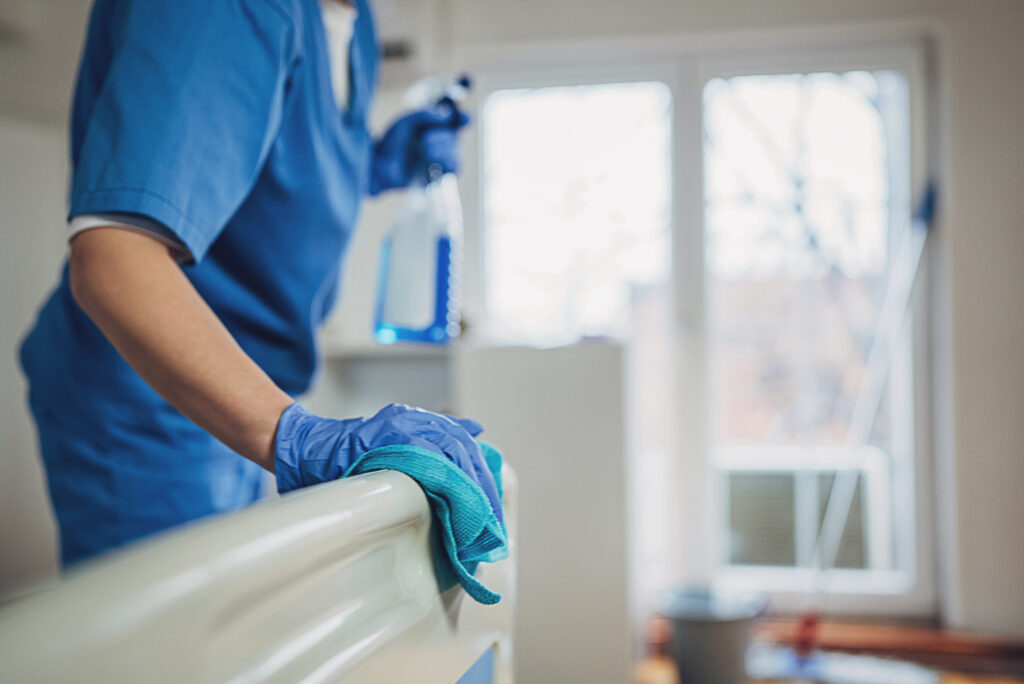 A person wearing blue scrubs and gloves cleans a hospital bed railing with a blue cloth and holds a spray bottle, standing near a sunlit window.