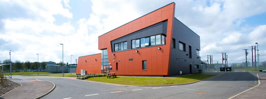 A modern, angular building with bright orange and black exterior panels, large windows, and security cameras sits beside a curved road under a partly cloudy sky. Grass and a picnic table are in front.