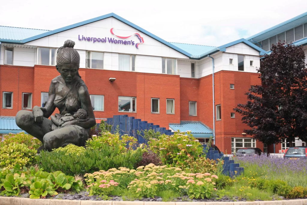 A statue of a mother holding a child sits in a garden in front of the Liverpool Women’s Hospital building, which has a red brick facade and blue roof accents.