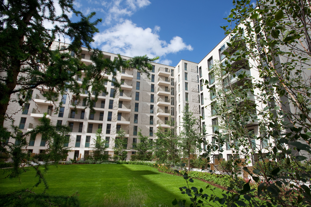 Modern apartment buildings with multiple balconies surround a landscaped courtyard with green grass, trees, and shrubs; the sky above is blue with scattered clouds.
