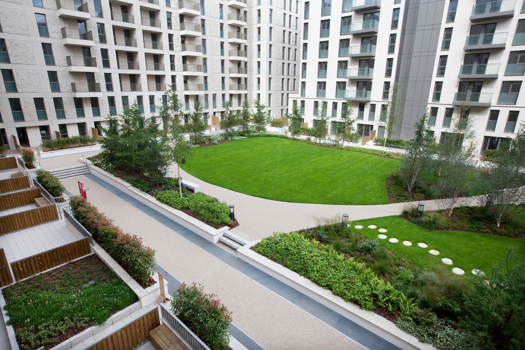 A modern apartment complex surrounds a landscaped courtyard with green grass, walking paths, neatly trimmed shrubs, and small trees. Balconies overlook the courtyard, and a few stone stepping circles decorate the lawn.