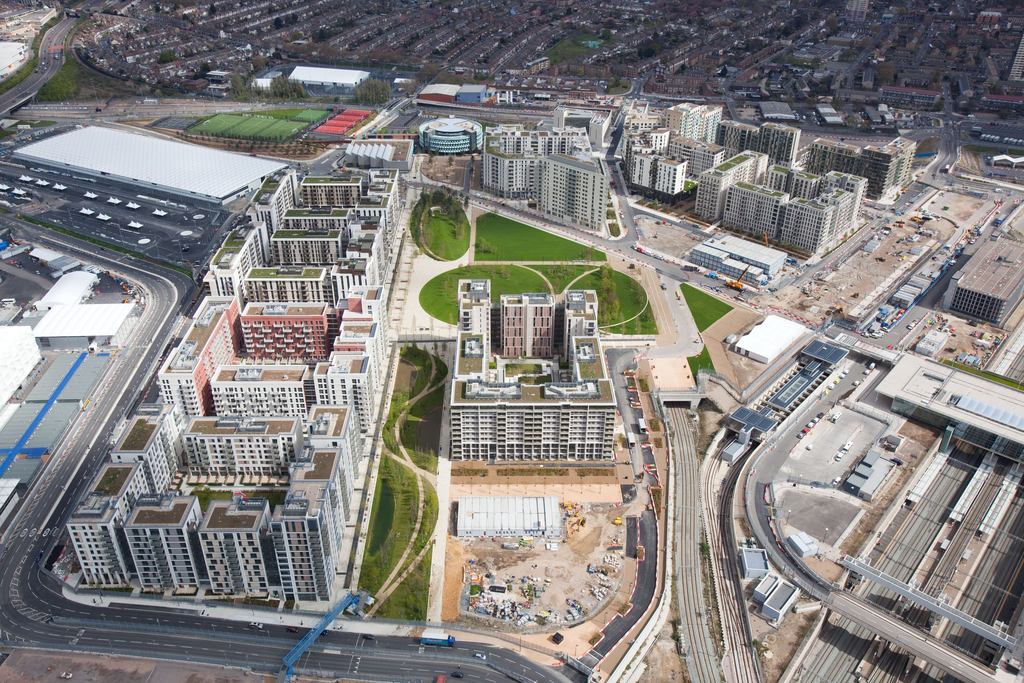 Aerial view of a modern urban development with multiple apartment buildings, green courtyards, roads, and ongoing construction, surrounded by a railway station and residential neighborhoods.