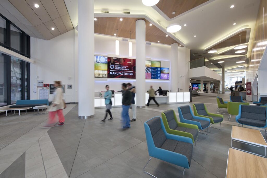 A modern, spacious lobby with bright lighting, colorful seating, several people walking, and a reception desk in the background beneath large digital screens displaying information.