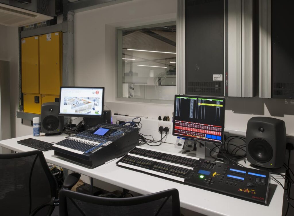 A modern audio-visual control room with computers, mixing consoles, speakers, and various equipment on a white desk, viewed through a window into a studio space.