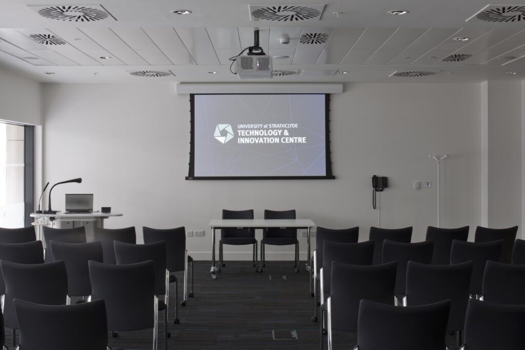 A modern conference room with black chairs facing a projector screen that reads University of Strathclyde Technology & Innovation Centre. The room has a lectern, tables, a projector, and wall-mounted equipment.