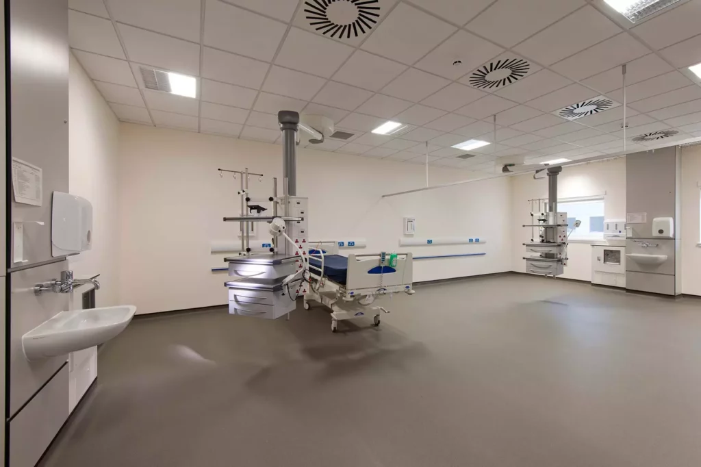 A modern, empty hospital room with a single hospital bed, medical equipment, sink, and clean white walls. The room is brightly lit with ceiling lights and has a sterile, uncluttered appearance.