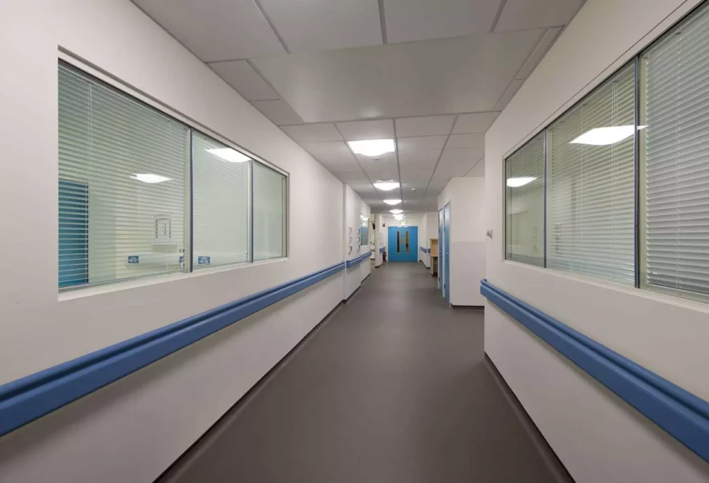 A clean, empty hospital corridor with blue handrails, white walls, large windows on both sides, and bright ceiling lights. Double doors are visible at the end of the hallway.