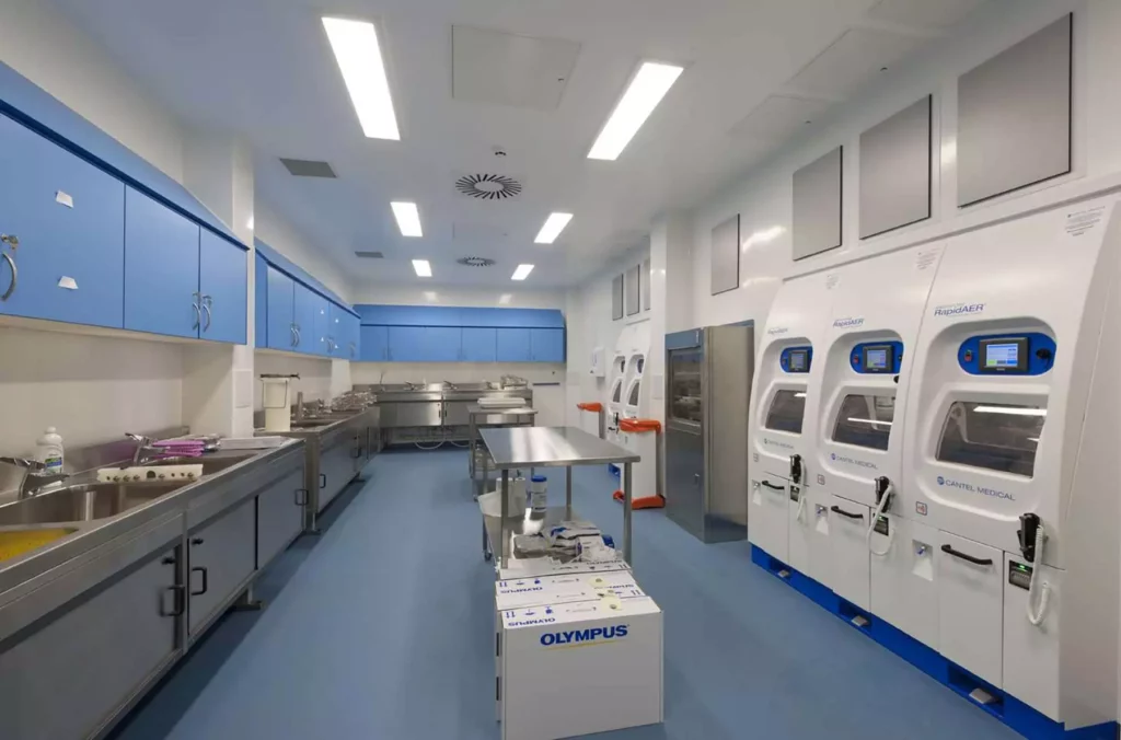 A modern, well-lit laboratory with blue cabinets, stainless steel counters, sterilization machines on the right, and a box labeled OLYMPUS on the blue floor in the center.