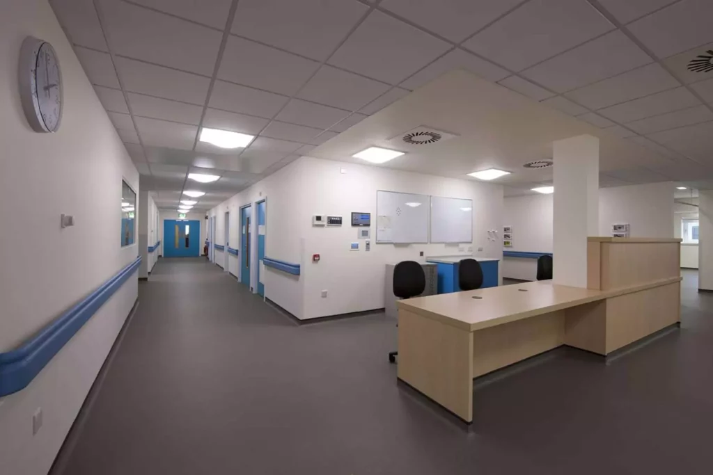 A clean, empty hospital corridor with blue accents, a reception desk, wall clock, and several doors leading to patient rooms. Bright ceiling lights illuminate the sterile, modern environment.