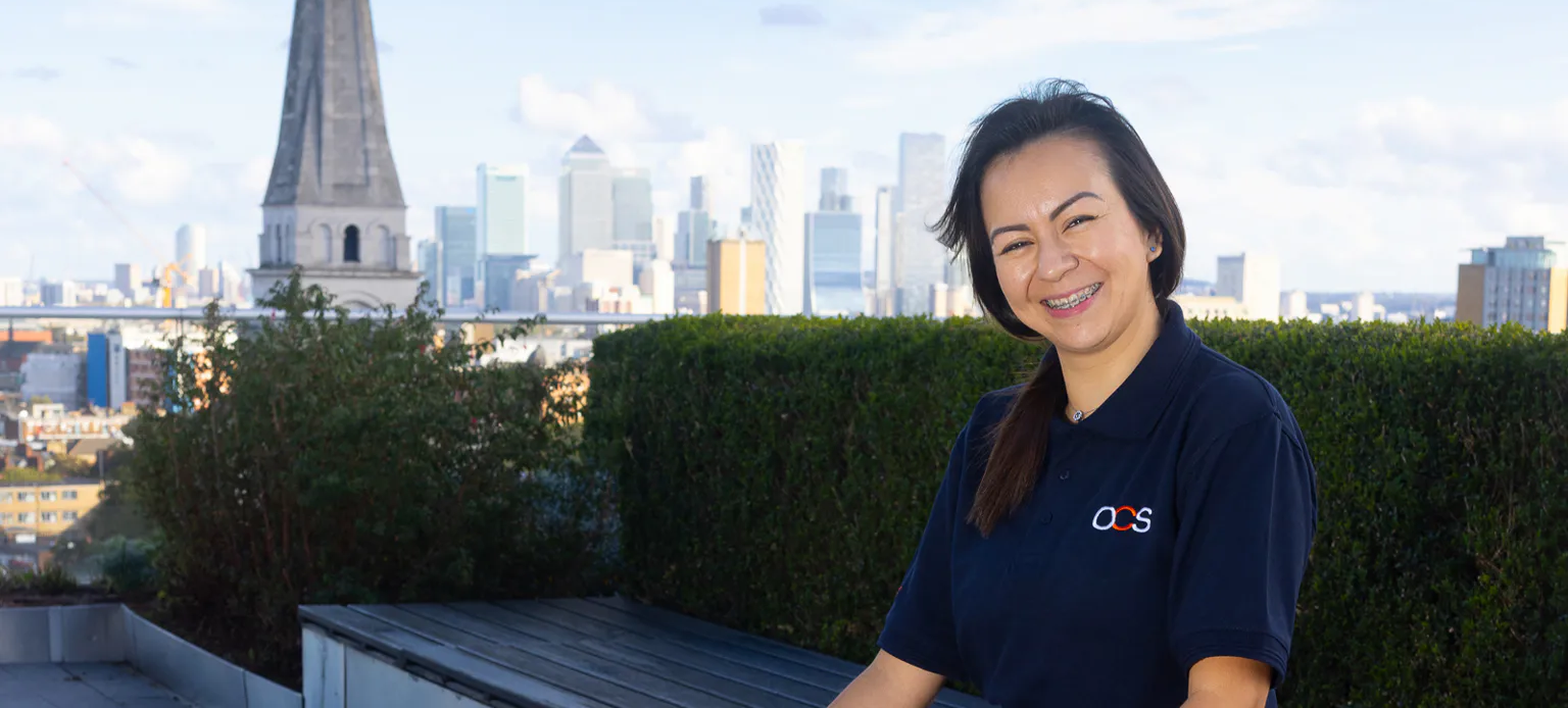 A woman in an OCS uniform smiles outdoors on a rooftop terrace, with a cityscape and skyscrapers in the background under a partly cloudy sky.