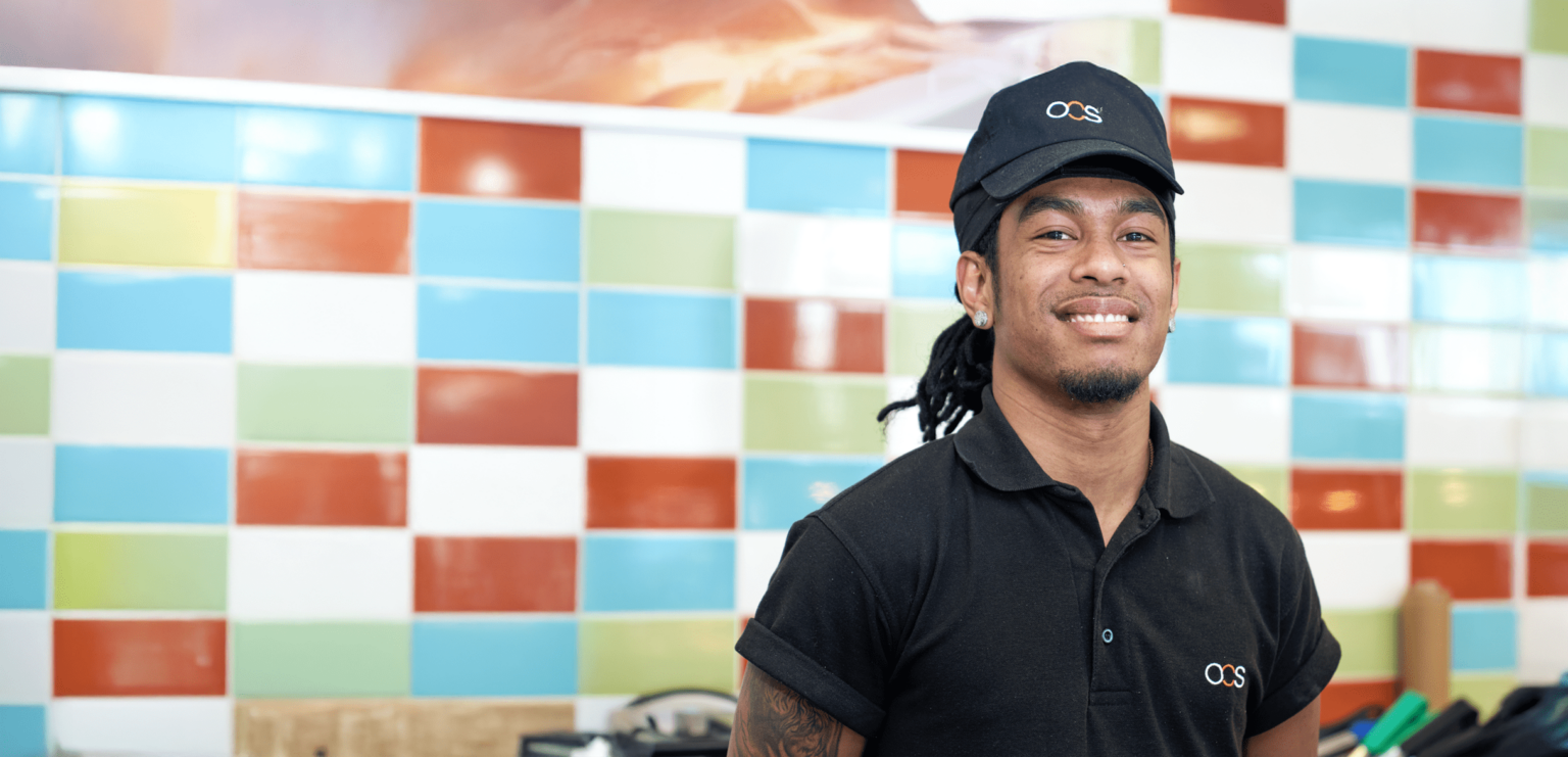 A smiling worker in a black polo shirt and cap with a white logo stands in front of a colorful tiled wall in what appears to be a restaurant or café.