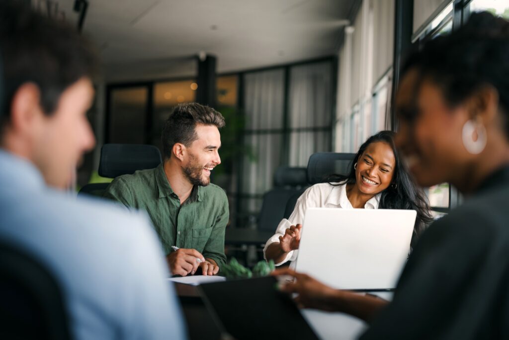Four people sit around a table in a modern office, two in focus smiling and talking while working on laptops. The atmosphere appears friendly and collaborative.