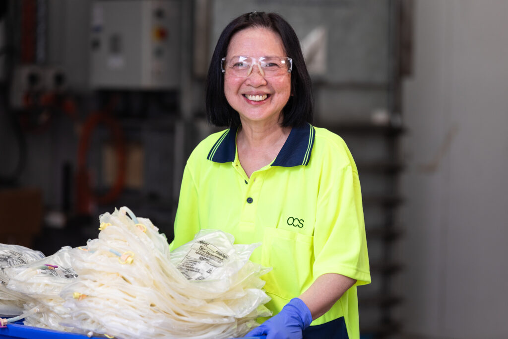 A woman wearing safety glasses and a bright yellow shirt smiles while handling a tray of packaged medical supplies in an industrial setting.