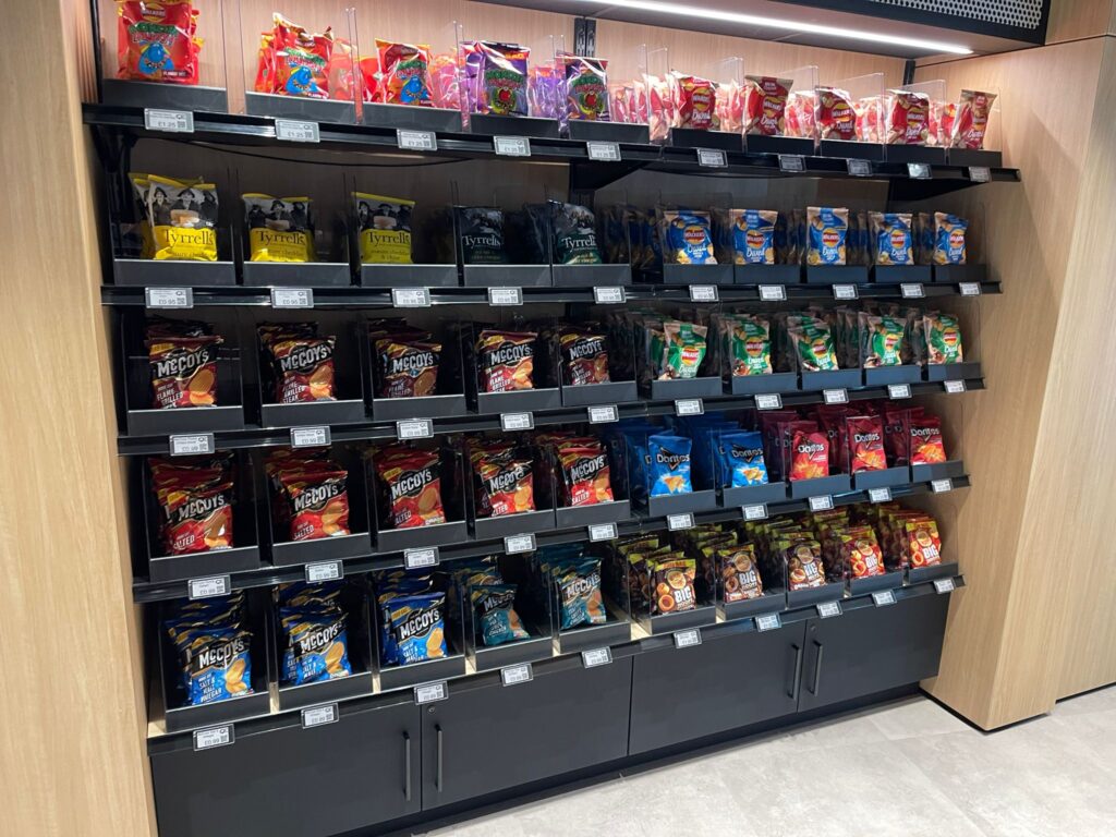 A supermarket shelf displays various brands and flavors of potato chips and snacks in colorful bags, organized neatly across five rows. The shelves are black with light wooden sides and electronic price tags.