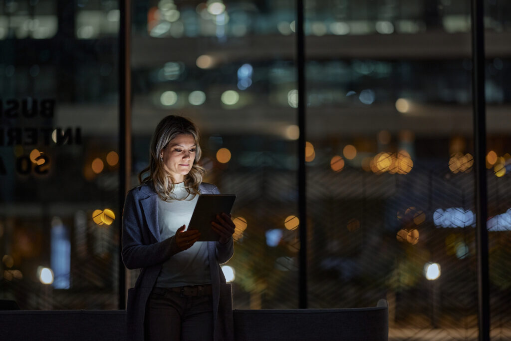 A woman stands indoors at night, illuminated by the light from a tablet she is holding, with large windows and city lights blurred in the background.