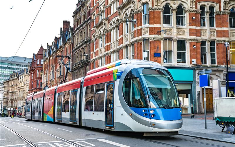 A modern light rail tram with a blue, red, and white design travels along tracks in a city center, surrounded by historic brick buildings with ornate architecture.