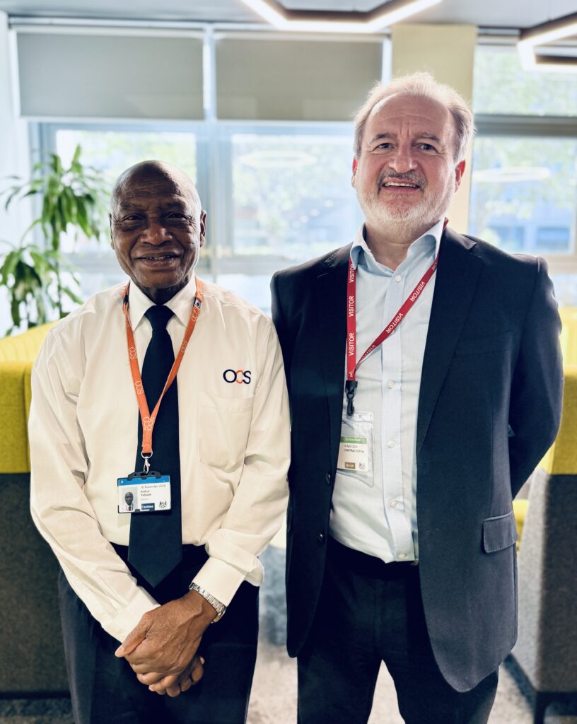 Two men wearing ID badges and lanyards stand side by side indoors, smiling at the camera. One wears a white shirt and tie, the other a dark suit. Bright office space with plants and large windows in the background.