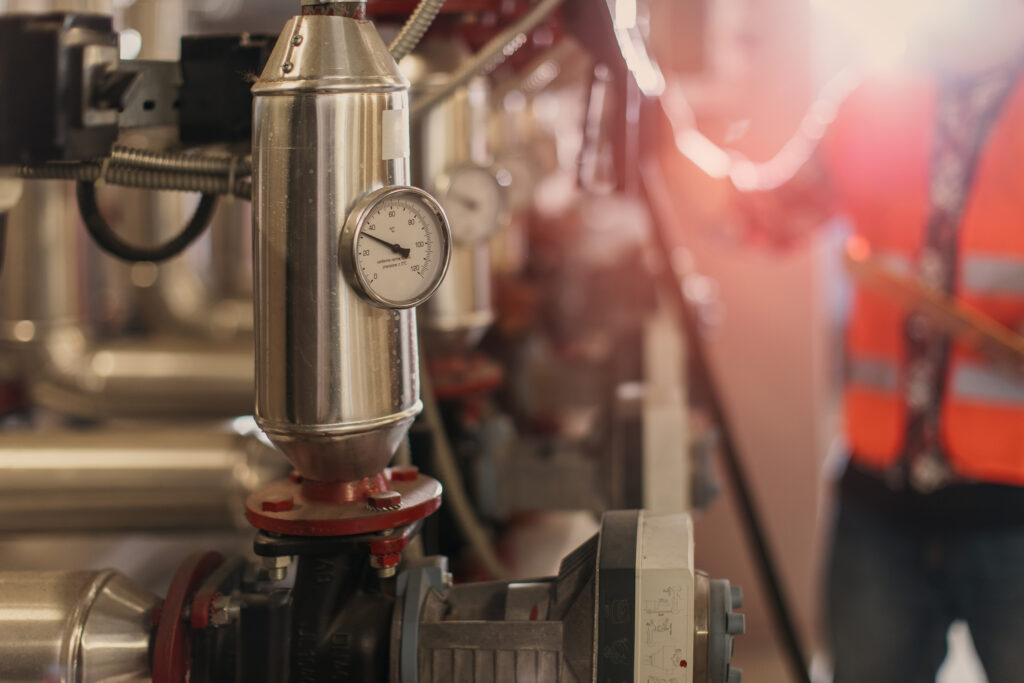 A close-up of industrial metal pipes with a pressure gauge attached, with a person in a high-visibility orange vest blurred in the background and sunlight shining into the scene.